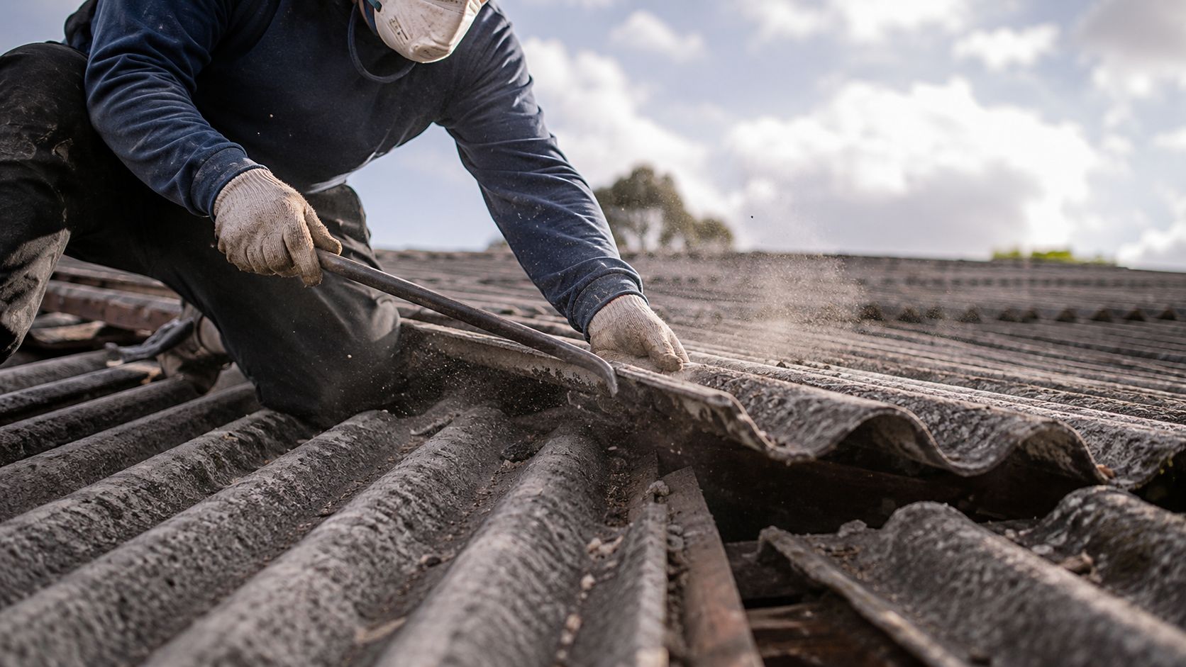 A construction worker wearing protective gear removing old corrugated asbestos roof panels with a crowbar.