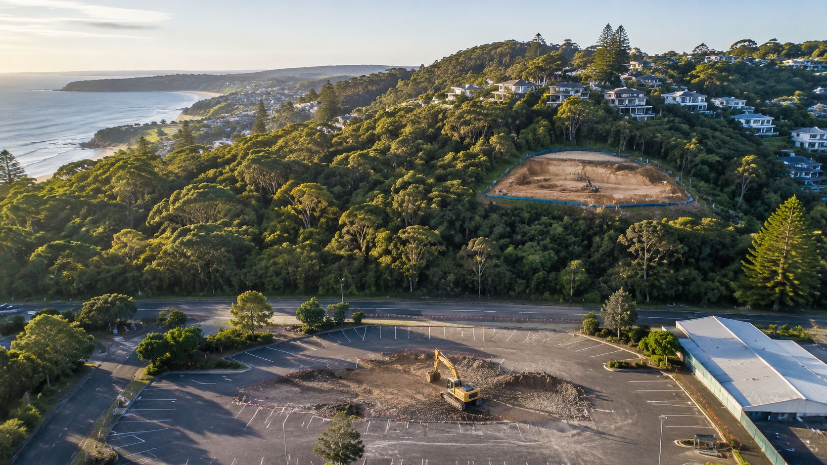 An aerial view showing an excavator at a construction site near a coastal town and forest.